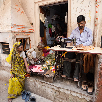 Varanasi, India - April 5, 2010: Indian tailor is using sewing machine on the street of Varanasi, India, while customer woman in sari is sitting by him.