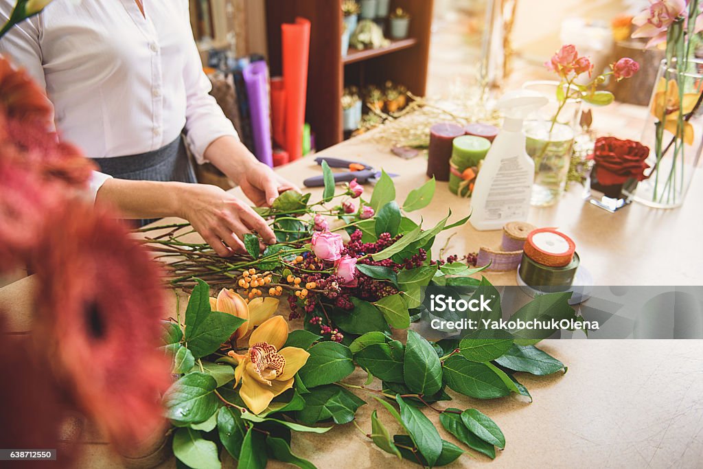 Professional florist creating masterpiece from plants Skillful woman is touching various flowers and making bouquet of it. Close up of her arms over the desk Desk Stock Photo