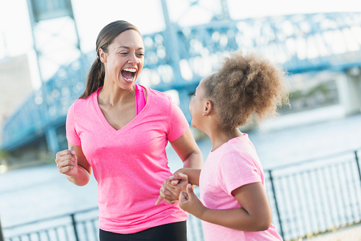 A black woman in her 30s standing outdoors by the waterfront in a city, laughing with her 7 year old daughter. They are wearing pink t-shirts, holding hands.