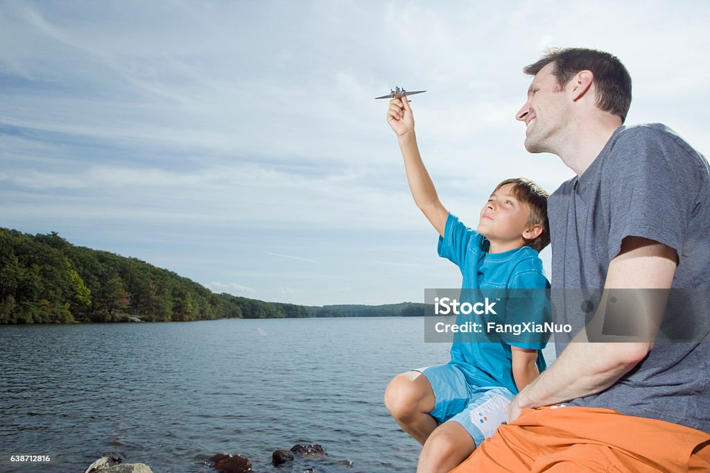 Father watching boy play with airplane next to lake Airplane Stock Photo