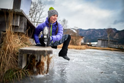 Woman with winter clothes and ice skate shoes sitting on the bench on the frozen lake and warming up. Cabins and hills are in the background.