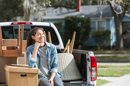 A young mixed race woman sitting on the back of a pickup truck loaded with boxes and furniture. She is moving, perhaps leaving home for the first time to go to college, or to move into her own apartment. She is dreaming about the future.