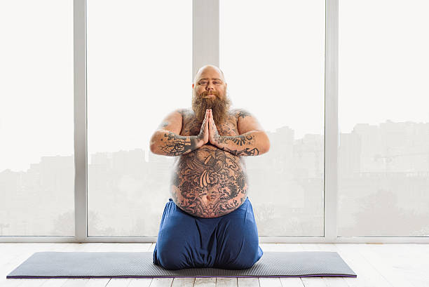 Serene fatso meditating in his apartment Calm mature fat man is doing yoga at home. He is sitting on mat and keeping hands together. Guy is looking at camera with satisfaction man yoga tattoo stock pictures, royalty-free photos & images