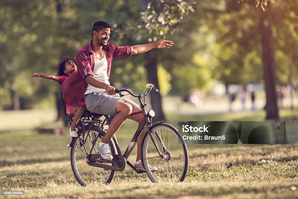 Carefree African American father and daughter cycling in the park. Happy African American father and daughter having fun together on a bicycle in nature. Cycling Stock Photo