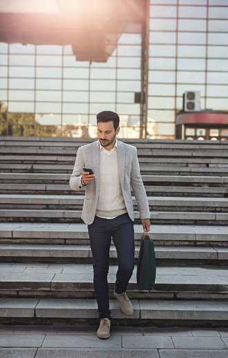 Young man in smart casual clothing carrying shopping bag and reading text message on the phone while going downstairs in the city.