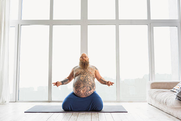 Relaxed thick guy meditating at home Serene fat man is doing meditation near window. He is sitting on mat. His bally is large with tattoo man yoga tattoo stock pictures, royalty-free photos & images