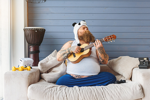 Cheerful male fatso is playing small guitar at home. His eyes are closed with pleasure