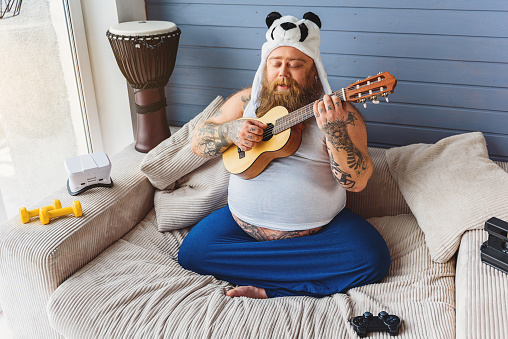 Carefree fat guy is singing to a guitar with inspiration. He is relaxing on sofa at home