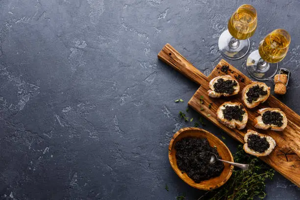 Sturgeon black caviar in wooden bowl, sandwiches and champagne on dark stone background copy space