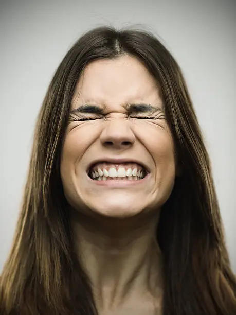 Close-up of angry young woman clenching teeth. Aggressive female is with eyes closed. She is against gray background. Vertical studio photography from a DSLR camera. Sharp focus on eyes.