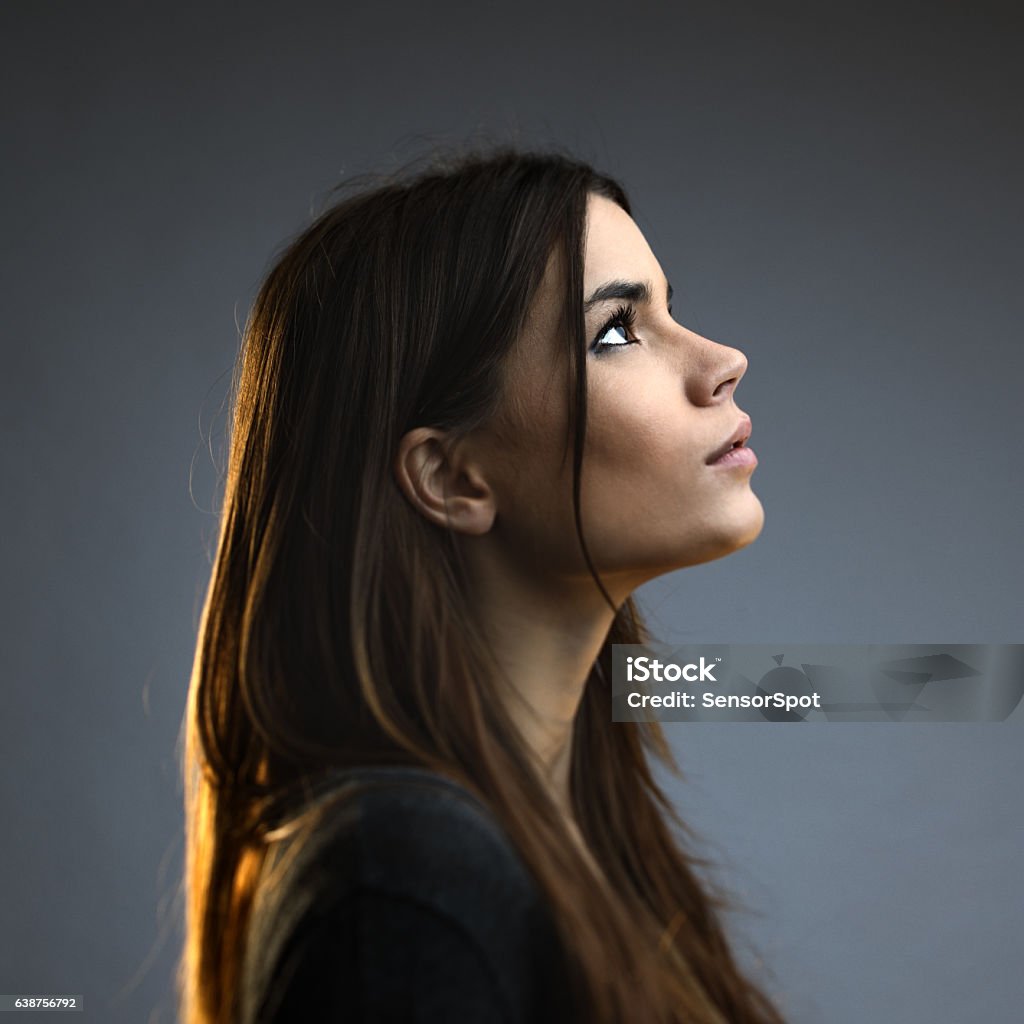 Beautiful woman posing against dark background Beautiful woman posing against against grey background. Beautiful female with long brown hair is standing. She is wearing casual clothes. Square studio photography from a DSLR camera. Sharp focus on eyes. Profile View Stock Photo