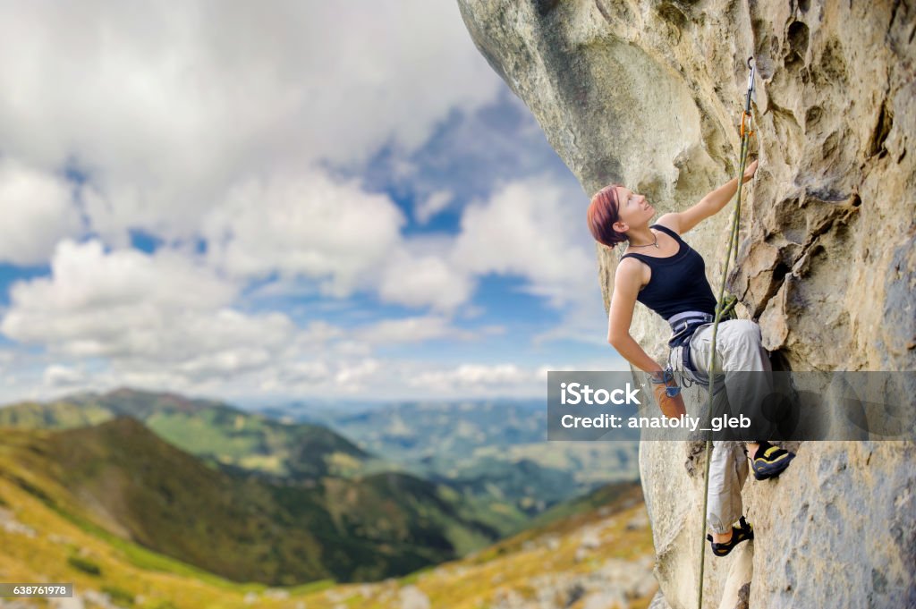 Female rock climber on steep overhanging rock cliff Young athletic female rock climber climbing cliff wall, hanging on one hand and holding hand in magnesium bag. Mountains and blue sky on the background. Copy space on the left. Mountain Climbing Stock Photo