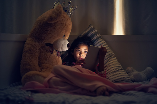 Shot of a little girl using a digital tablet while lying in bed with her teddy at night
