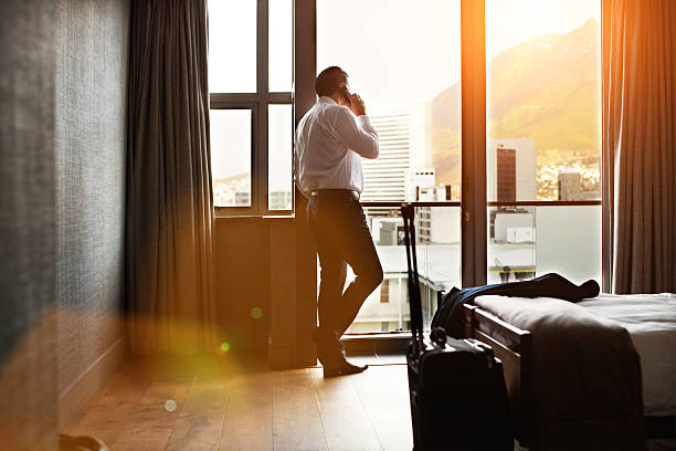 It was a long day but I'm back at the hotel Rearview shot of a businessman looking through a hotel room window and talking on a cellphone business travel stock pictures, royalty-free photos & images