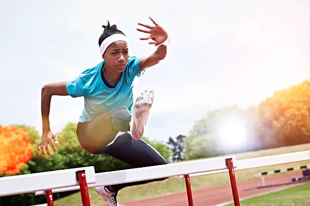 Shot of a young woman in mid air while jumping over a hurdle on a sports field