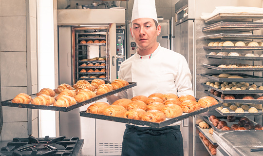 Italian chef with his freshly baked croissants. The young man is taking out from the oven two baking sheets full of croissants.