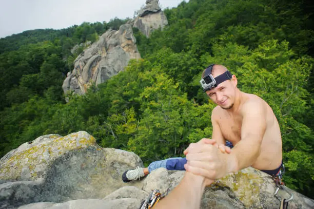 Young shirtless male climber is reaching for a helping-hand from his partner at the top of his climbing route in nature at the mountains. Action camera is attached to the man's head. Top view.