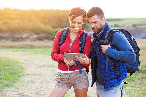 Young people using digital tablet for navigation in hiking. One girl and one man with backpacks and sweatshirts hiking on mountain. Nature view and sunlight on background.