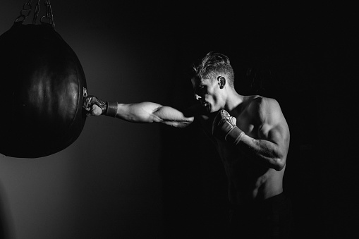 Image of a athletic young adult man punching bags with boxing gloves on. Horizontal composition, with backlight toned to black and white in post processing. This young man is working out, and this image could be used in many athletic sport types of media. Staying physically active is an important part to staying healthy. This young man takes care of his body by giving it the exercise it deserves. 