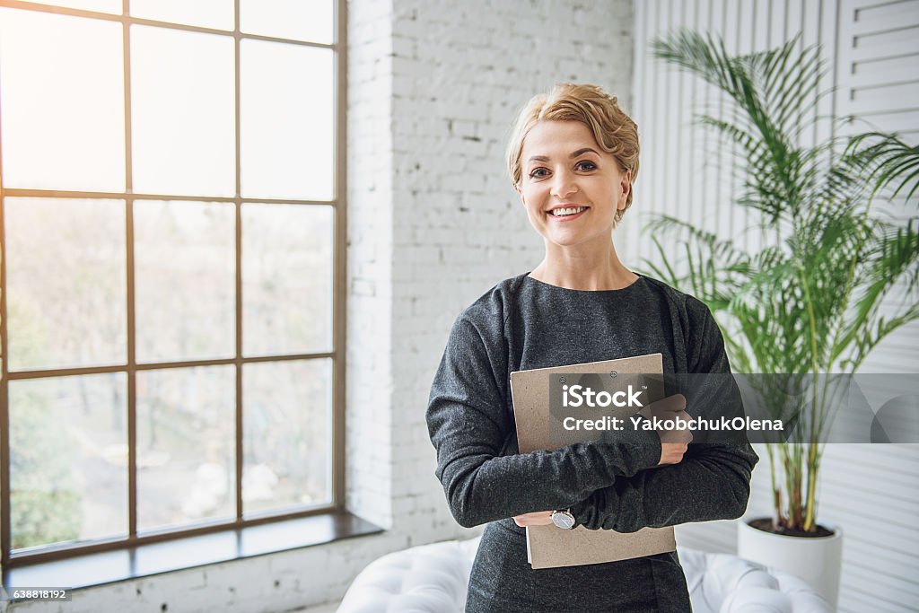 Sincerely smiling woman holding tablet Happy consulter is standing in front of window with bright smile on her face. She keeping folder of papers Digital Tablet Stock Photo