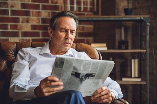 Senior concentrated businessman is sitting on sofa. He looking through newspaper articles