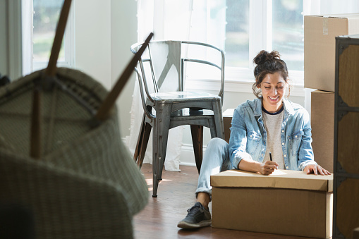 A young mixed race woman, Hispanic and Pacific Islander ethnicity, moving into a house or apartment. She is sitting on the floor writing a note or going through a checklist.