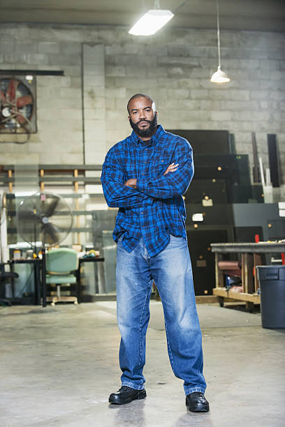 African American man in a factory workshop An African American man in his 30s looking at the camera with a serious expression, arms folded. He is a worker in a factory workshop. man beard plaid shirt stock pictures, royalty-free photos & images