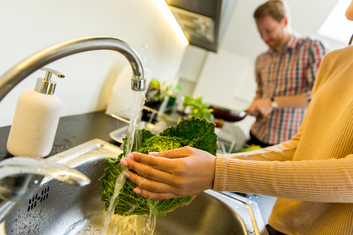 Young woman washes kale on the tap while a young man in the background cooking
