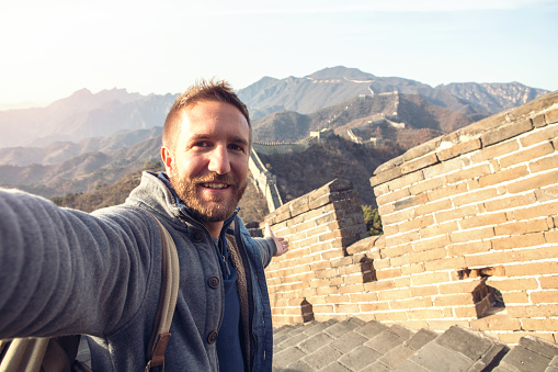 Young man on top of the Great Wall of China taking a selfie portrait with the beautiful landscape from high up. View of the wall going on the mountains for kilometres, majestic scenery of a winter day.