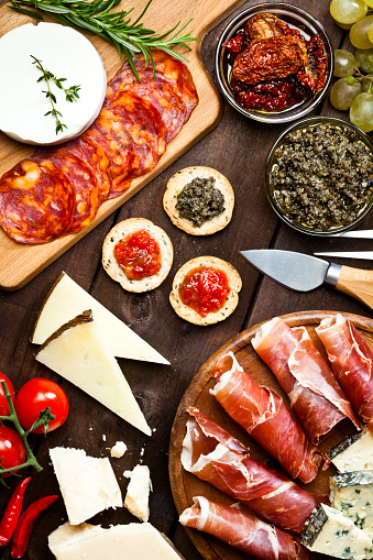 Top view of a rustic wood table filled with a assortment of fine food. A wood tray with prosciutto and cheese is at the bottom-right of the frame. Two glass bowls with dried tomatoes and olives paste and a cutting board are at the top. Various types of cheese complete de composition. DSRL studio photo taken with Canon EOS 5D Mk II and Canon EF 100mm f/2.8L Macro IS USM