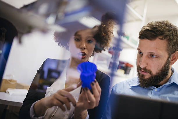 Amazed by 3D printing Close up shot of 3D printer printing 3D objects. Two workers are observing the process. Women is holding 3D object in her hand. polymer photos stock pictures, royalty-free photos & images