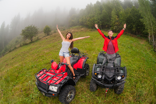 Happy beautiful couple standing on four-wheelers ATV smiling and holding thier hands up. Wide view from high piont