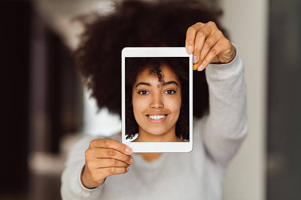 Mixed race woman taking selfie with tablet Smiling girl showing photo on tablet in front of photos stock pictures, royalty-free photos & images