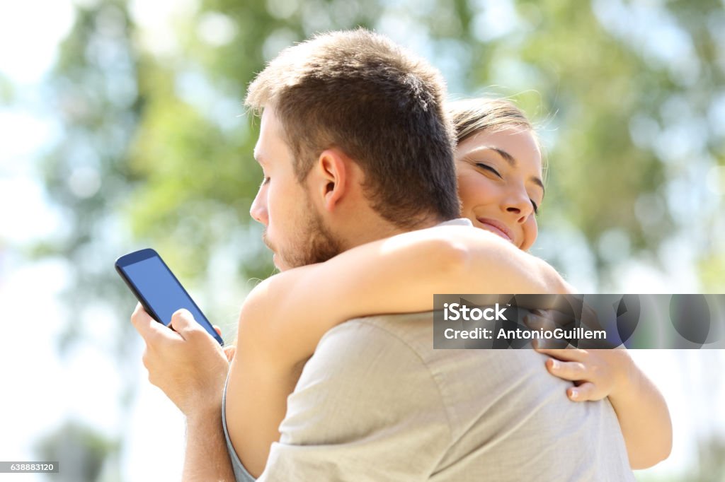 Cheater hugging his innocent girlfriend Cheater texting with his other lover on phone and hugging his innocent girlfriend Infidelity Stock Photo