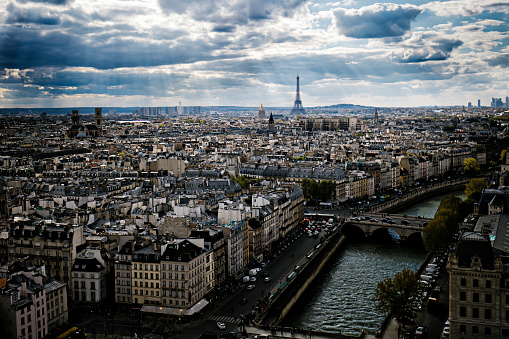 This image depicts a high-altitude view of a dense urban cityscape. The vantage point suggests it could be from a high floor of a tower or a skyscraper. The city is sprawling with numerous buildings packed tightly together, with their rooftops forming a somewhat uniform grey sea of structures. The architecture appears to be consistent with that of a European city, possibly Paris, given the buildings' style and density. In the distance, there are taller high-rise buildings that seem more modern, possibly marking the city's outer neighborhoods or a business district. The photograph is taken during the daytime under a clear sky with a few scattered clouds, allowing for a clear view of the expansive city below.