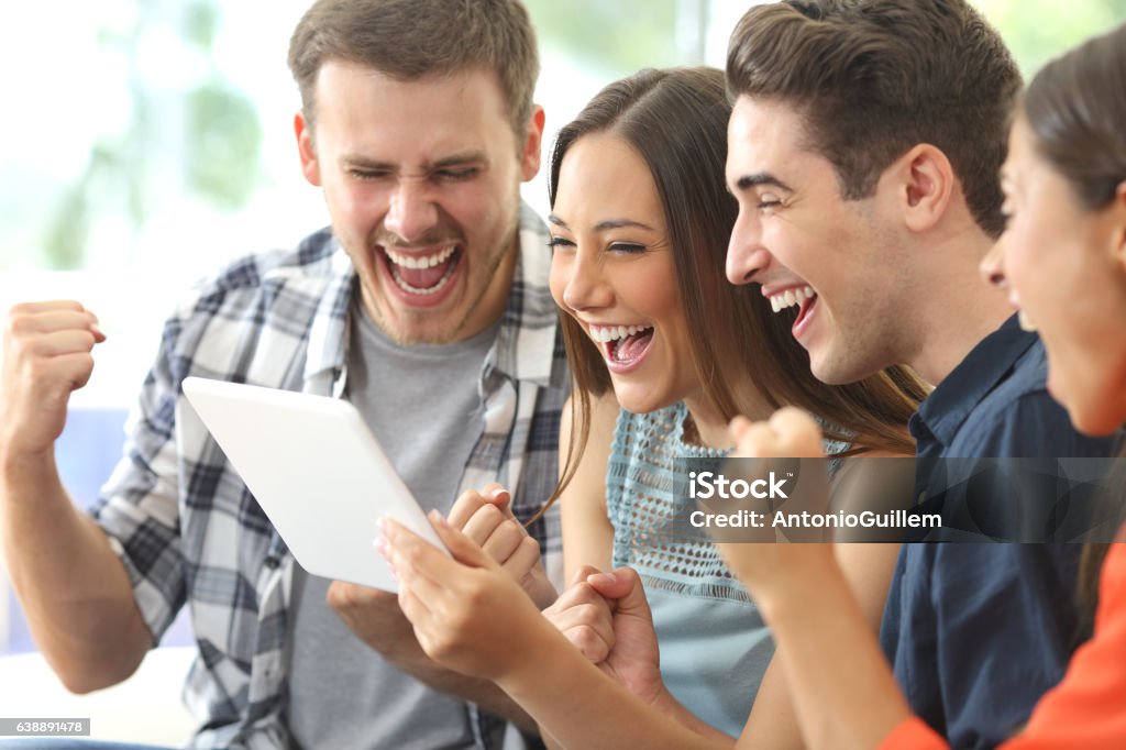 Excited group of friends watching tv from tablet Excited group of four friends viewing media content on line from a tablet in a house interior Educational Exam Stock Photo