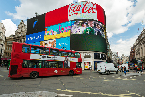 The iconic Regent LCD screens with tourists and a red bus passing by in Piccadilly Circus late in the day.