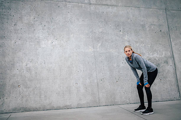 Woman athlete runner taking a break Full length shot of young beautiful female leaning on gray background while resting after everyday training. Woman athlete runner taking a break with copy space. hand on knee stock pictures, royalty-free photos & images