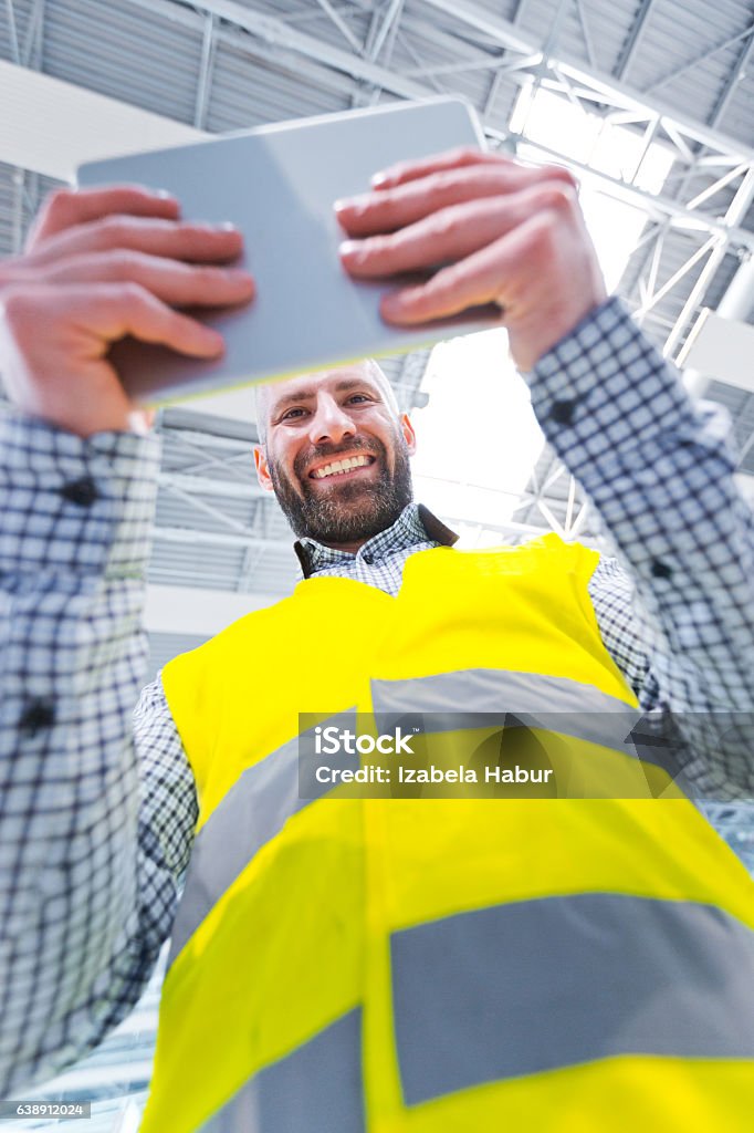 Engineer using a digital tablet, low angle view Engineer using a digital tablet in a industrial hall, smiling at camera, low angle view. Adult Stock Photo