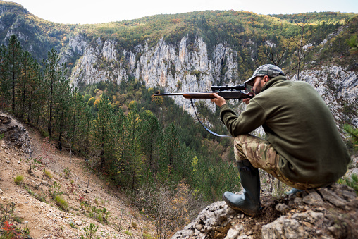 Portrait of hunter with sniper rifle while crouching on top of the hill and aiming at the target.