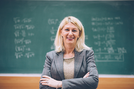 Portrait of mature academic person, female professor standing near blackboard with math forumlas. Woman is 50-60 years old, with blonde hair and sophisticated appearance. Image taken in Europe.