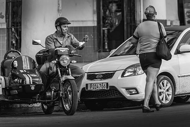 Havana, Cuba- October 11, 2016. Wide Havana boulevard exterior. Cuban woman crossing the road while the man on vintage motorcycle is looking at her.. She is wearing summer attire and sun glasses. Typical city scene win Havana streets.