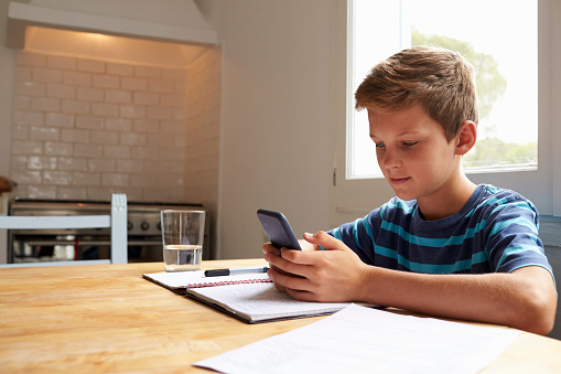 Boy Uses Mobile Phone Whilst Doing Homework At Kitchen Table