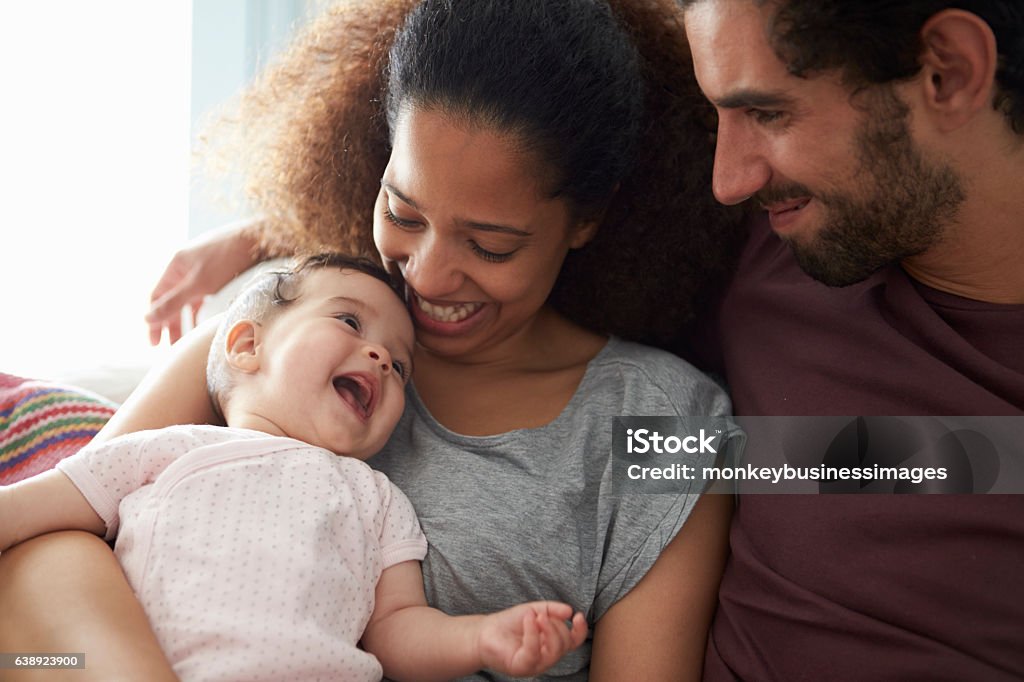 Parents Sitting On Sofa Cuddling Baby Daughter At Home Baby - Human Age Stock Photo