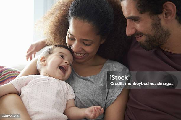 Parents Sitting On Sofa Cuddling Baby Daughter At Home Stock Photo - Download Image Now