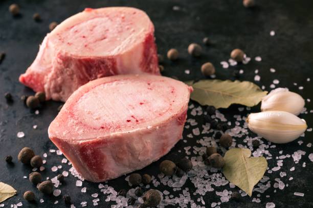 Detail of two slices of marrowbone on black board Horizontal photo with two pieces or slices of big marrowbone with few drops of blood and meat on side. Bones are placed on worn black baking tray with salt and other spices around. pink pepper spice ingredient stock pictures, royalty-free photos & images