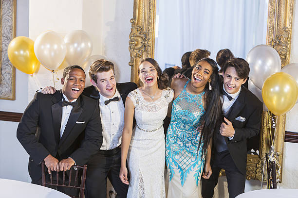Teenagers and young adults in formalwear at party A group of five multi-ethnic teenagers and  young adults dressed in formalwear - dresses and tuxedos. They are at a special event, a party or prom, standing together in a row, laughing and smiling. cocktail dress stock pictures, royalty-free photos & images