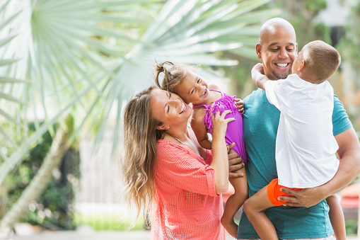 A happy mixed race family with two children on a tropical vacation standing outdoors. The father is holding the 6 year old boy and the mother is holding the 2 year old girl. They are African American and Caucasian.