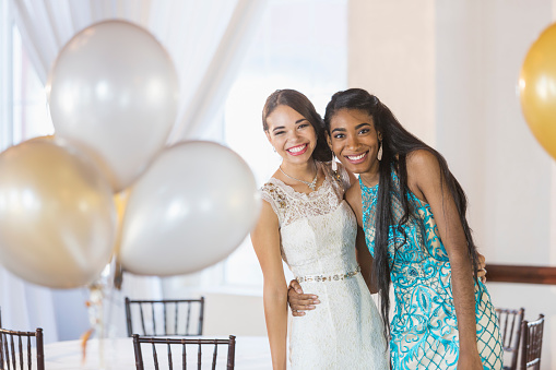 Two multi-ethnic young women wearing dresses, posing together in a party room in a restaurant, surrounded by balloons, smiling at the camera.