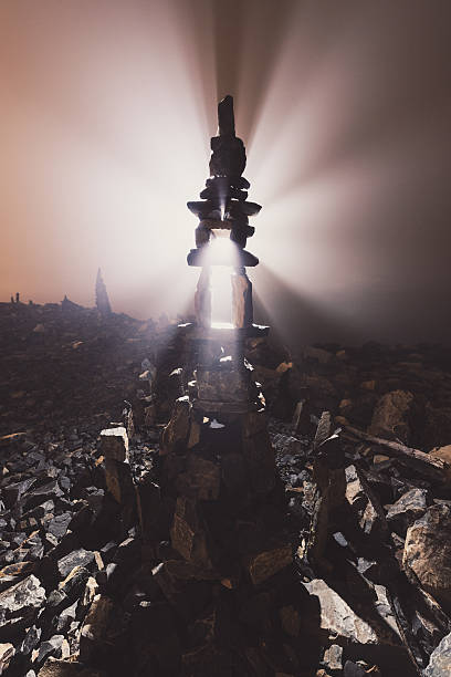 A tall inuksuk is backlit in heavy fog, rays of light emanating outward.  Long exposure.